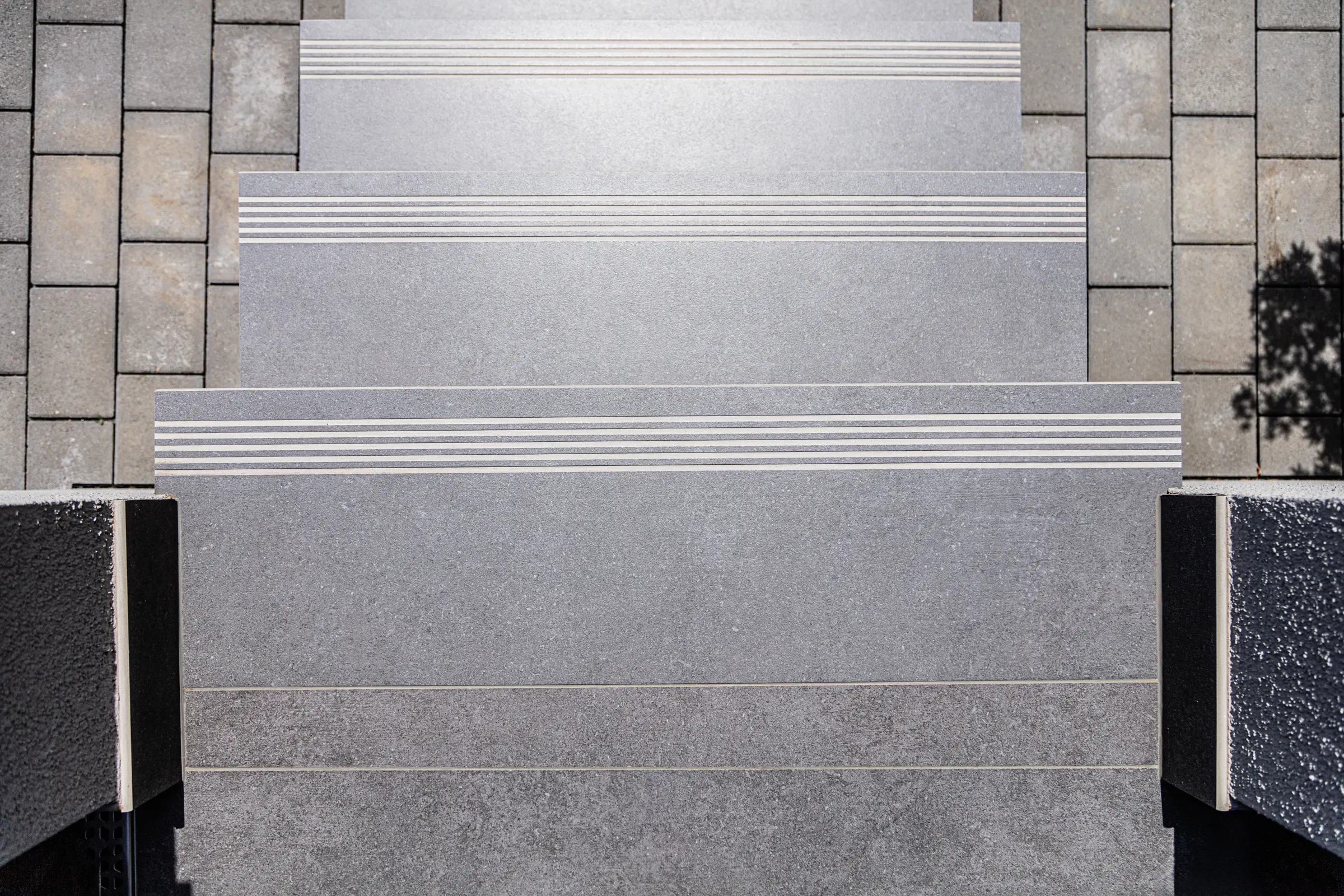 Draufsicht auf graue geflieste Außentreppe mit Rutschhemmung und angrenzendem Pflaster bei Sonnenlicht