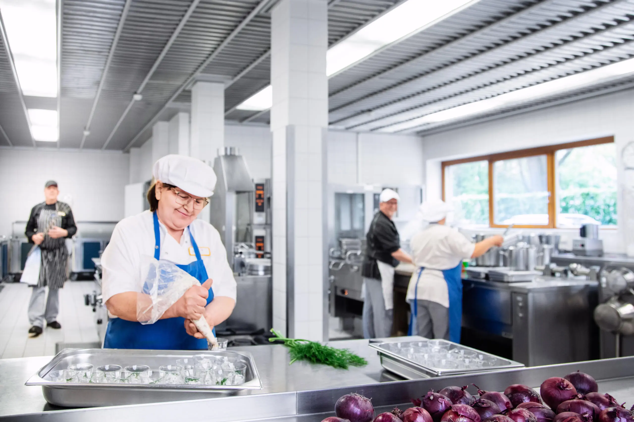Köchin in Arbeitskleidung beim Anrichten von Speisen in einer hellen Großküche, im Vordergrund rote Zwiebeln und Dill, im Hintergrund weitere Mitarbeiter