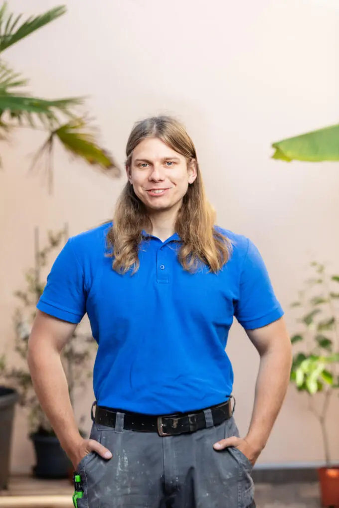 Lächelnder Handwerker mit langen blonden Haaren in blauem Poloshirt und grauer Arbeitshose vor beigem Hintergrund mit Pflanzen