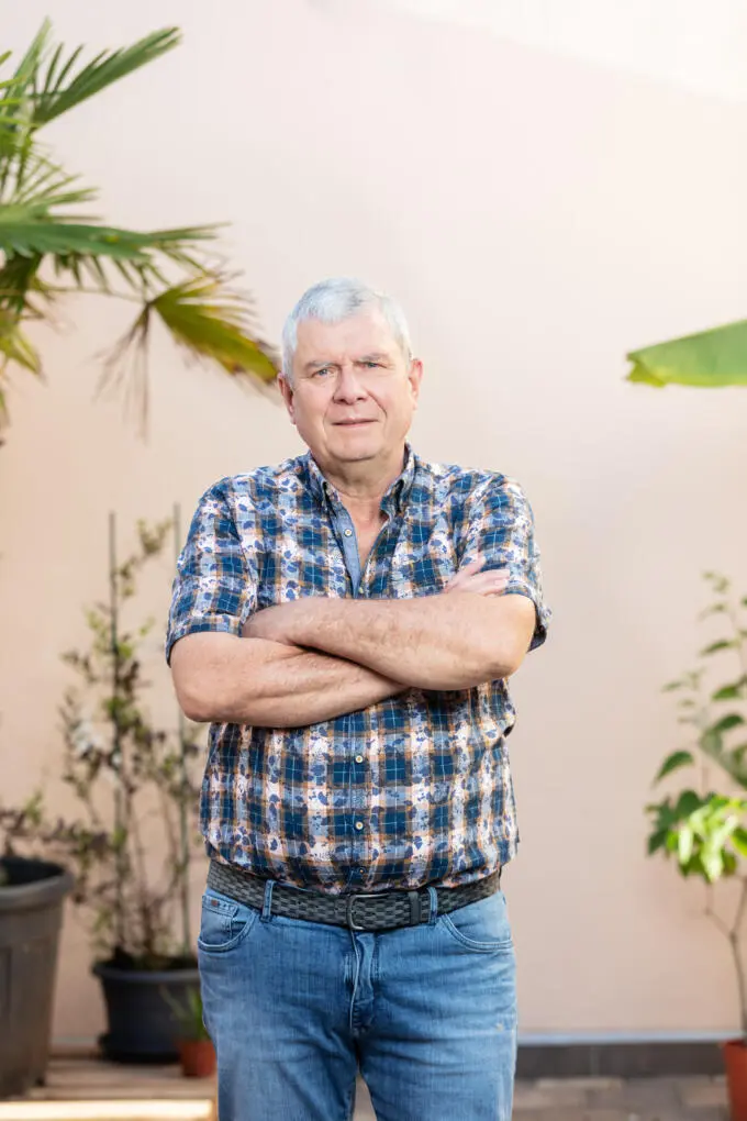Älterer Mann mit grauen Haaren und verschränkten Armen in kariertem Hemd und Jeans vor beiger Wand mit Pflanzen