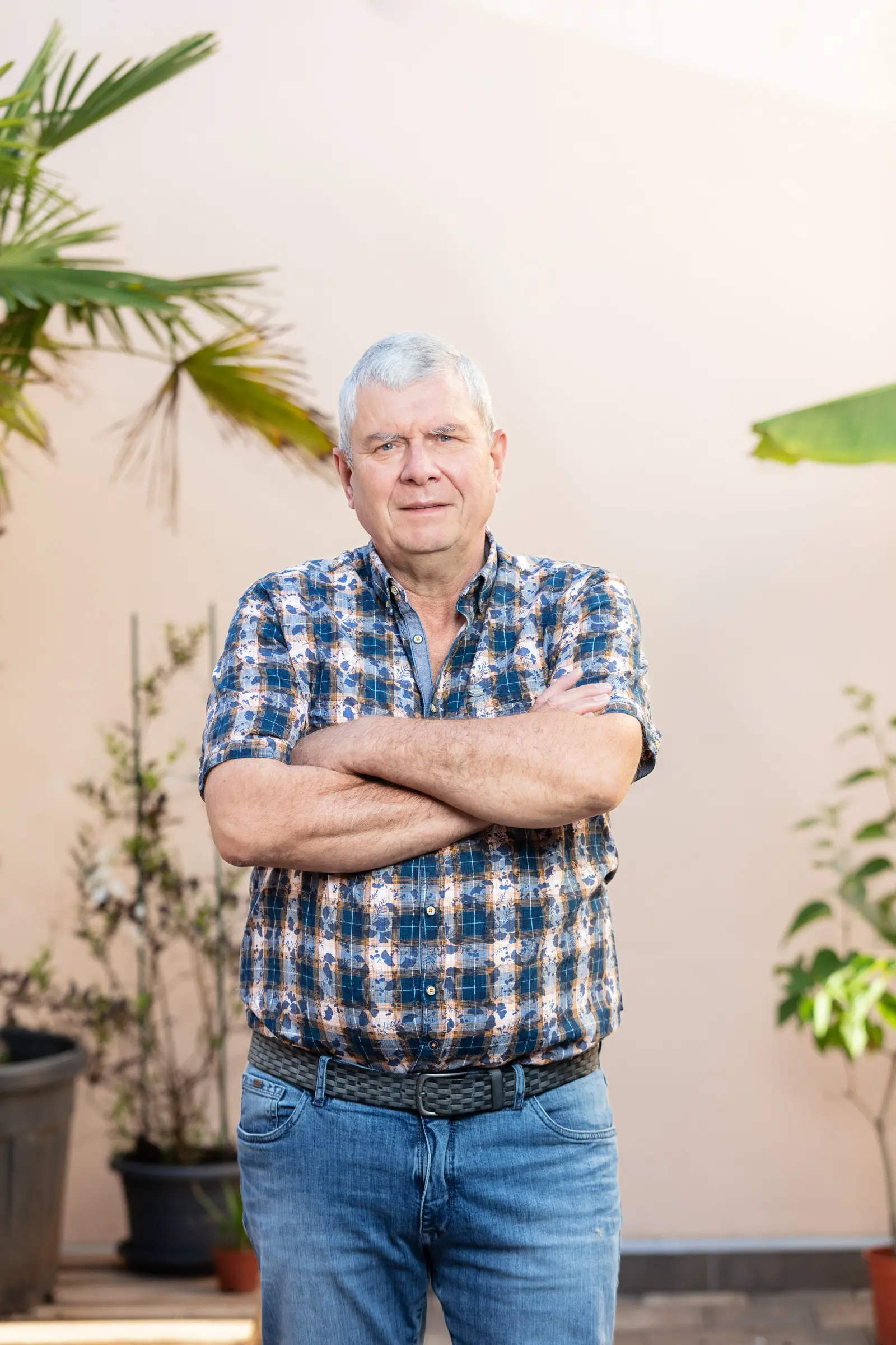 Älterer Mann mit grauen Haaren und verschränkten Armen in kariertem Hemd und Jeans vor beiger Wand mit Pflanzen
