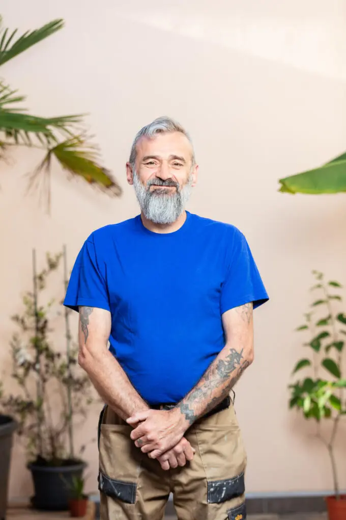 Porträt eines lächelnden Handwerkers mit grauem Bart und blauem T-Shirt vor beiger Wand mit Pflanzen