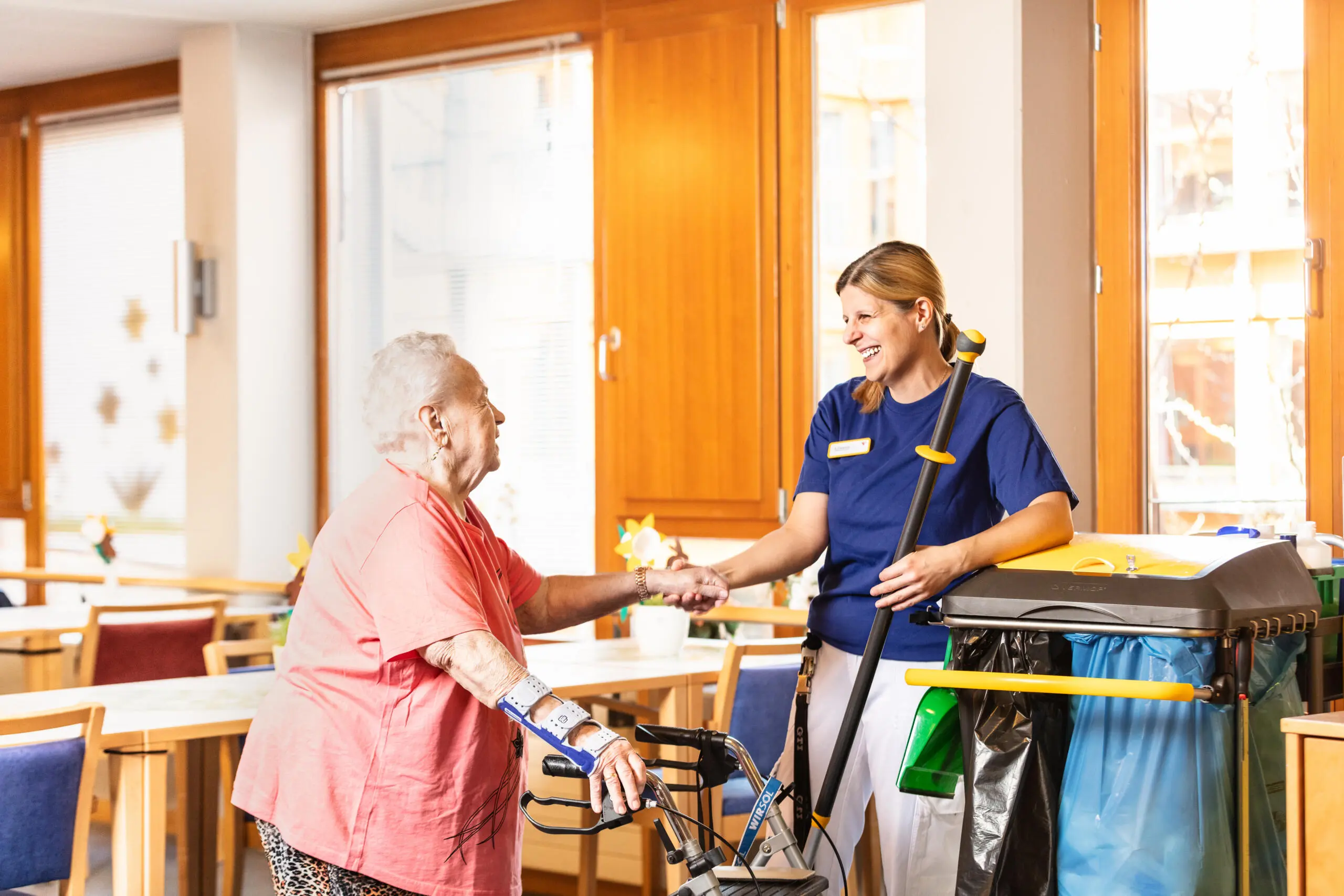 Freundliche Pflegekraft schüttelt Hand einer älteren Dame mit Rollator im hellen Raum.