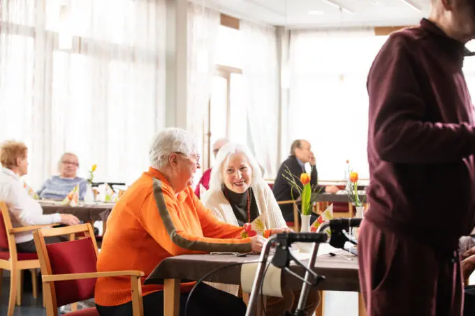 Ältere Menschen sitzen an Tischen in einem hellen Raum, angeregte Unterhaltung.