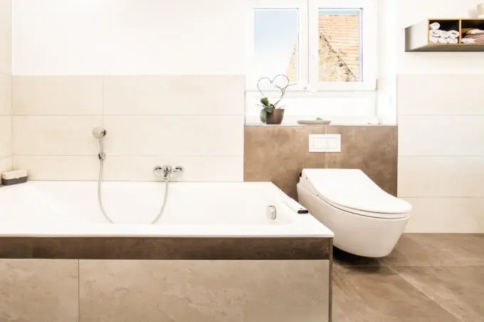 Modernes Badezimmer mit beigen Wandfliesen, braunen Akzentfliesen, weißer Badewanne und Hänge-WC unter einem Fenster