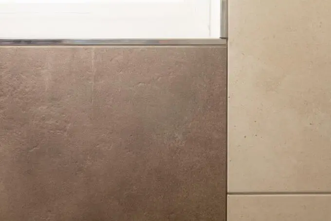 Detailaufnahme eines Fliesenabschlusses mit Metallprofil zwischen dunkler Fliese und weißem Fensterrahmen neben Naturstein