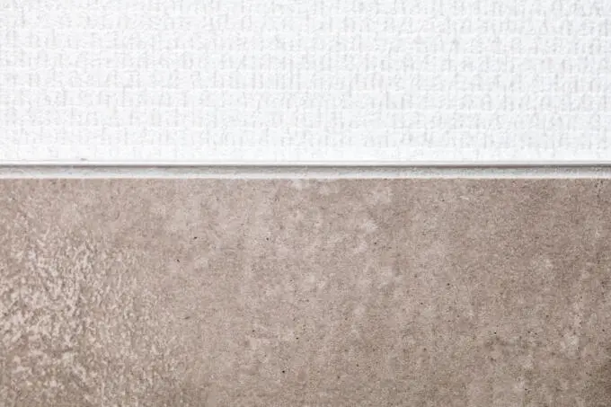 Detailaufnahme eines horizontalen Materialübergangs zwischen weißer Struktur-Wandfläche und beiger Fliese mit Trennprofil