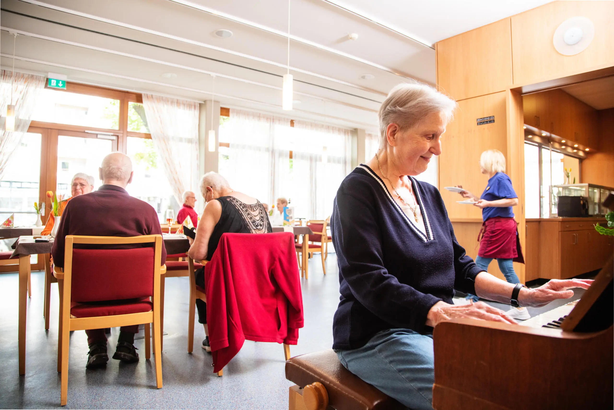 Frau spielt Klavier in hellem Raum mit Senioren an Tischen.