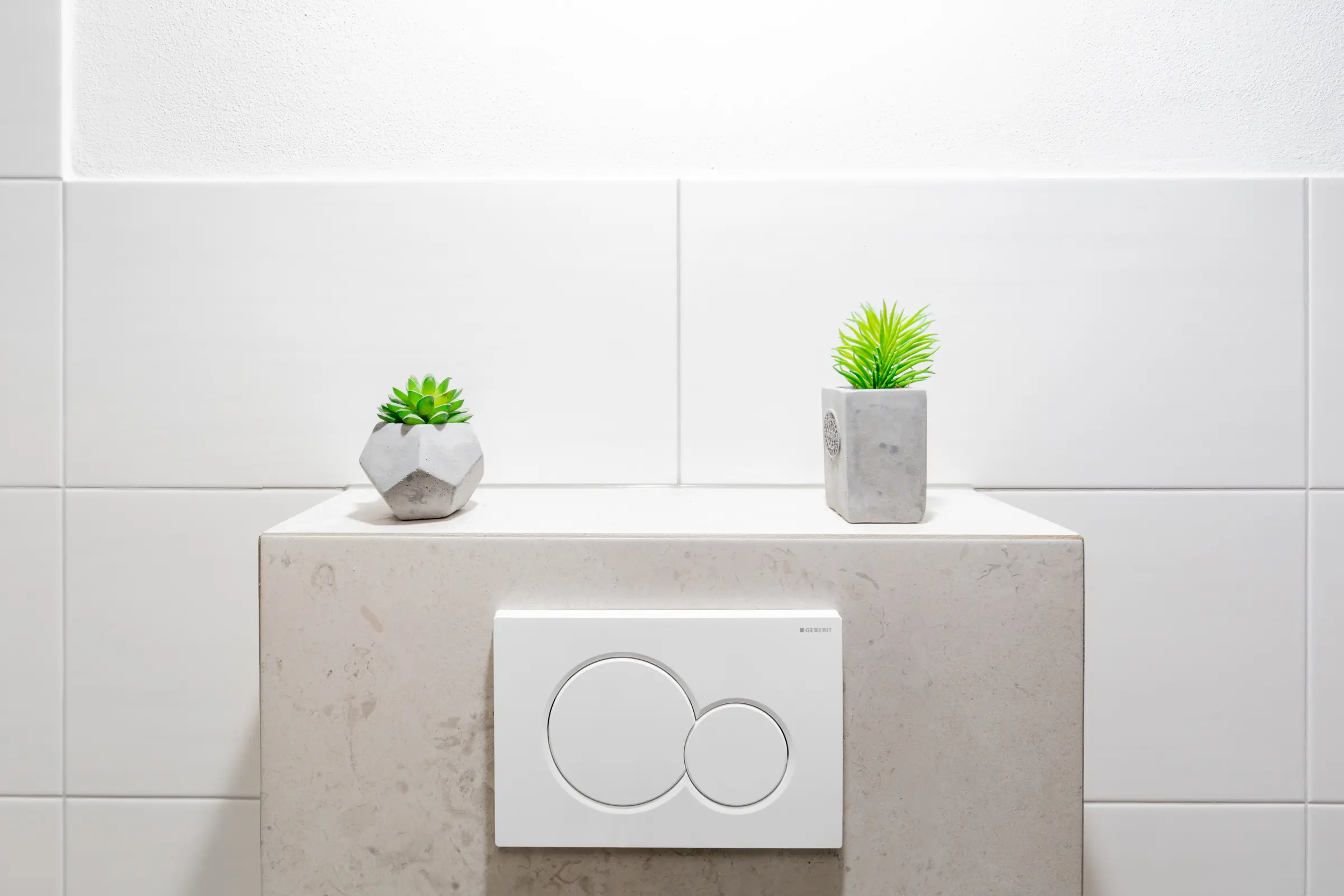 Detailaufnahme einer gefliesten WC-Vorwandinstallation mit weißer Betätigungsplatte und Pflanzendekoration vor weißen Wandfliesen