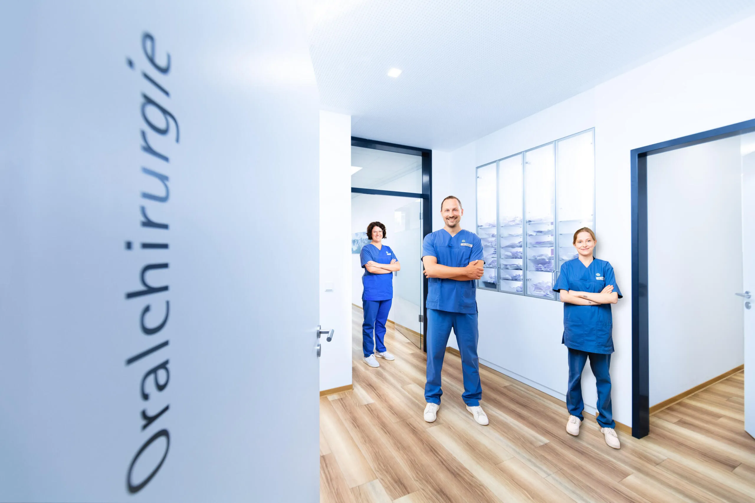 Drei medizinische Fachkräfte in blauer Berufsbekleidung stehen mit verschränkten Armen lächelnd in einem hellen Praxisflur, im Vordergrund unscharfer Schriftzug Oralchirurgie.