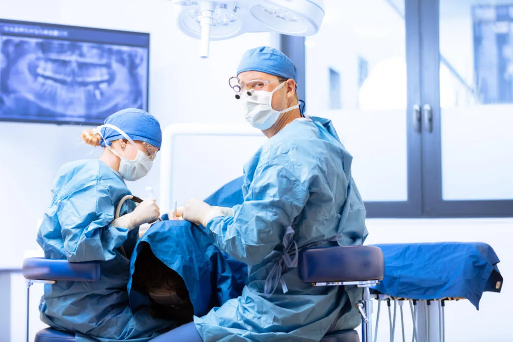 Ein Zahnchirurg mit Lupenbrille und eine Assistentin in blauer Schutzkleidung führen einen Eingriff an einem Patienten durch, im Hintergrund ist ein Röntgenbild auf einem Monitor sichtbar.