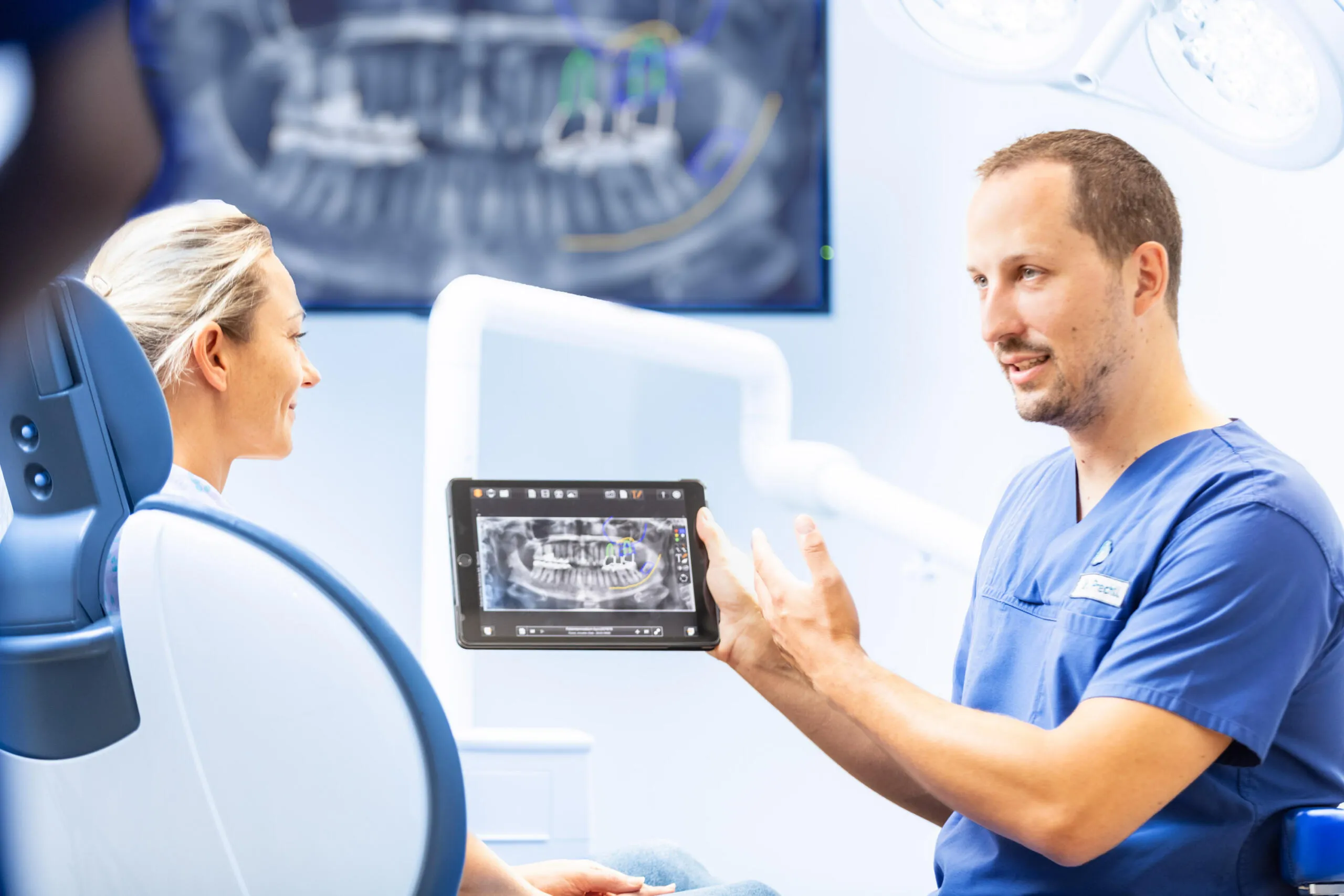 Ein Zahnarzt in blauer Berufskleidung zeigt einer Patientin auf dem Behandlungsstuhl ein digitales Röntgenbild auf einem Tablet.