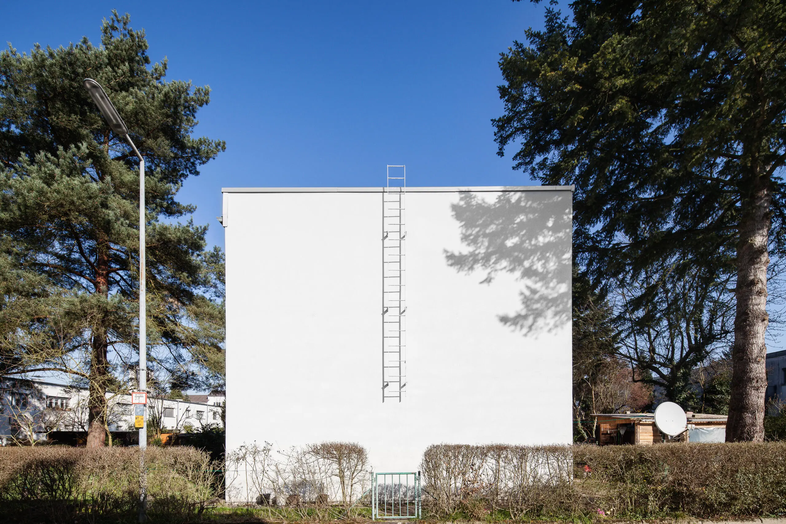 Eine weiße, fensterlose Gebäudefassade mit einer zentralen Metallleiter, eingerahmt von Bäumen unter blauem Himmel.