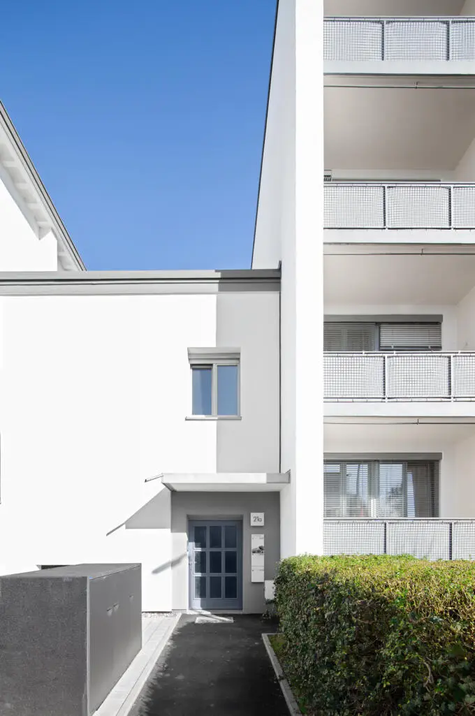 Außenansicht eines weiß verputzten Wohngebäudes mit grauer Eingangstür, Hausnummer 21a und seitlichen Balkonen mit Metallgittergeländern bei blauem Himmel.