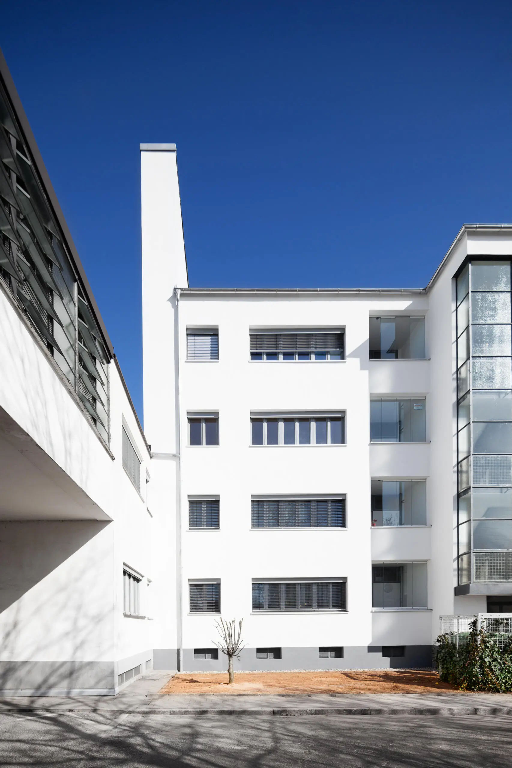 Außenansicht eines weißen Gebäudekomplexes der klassischen Moderne mit geometrischer Fassade und vertikalen Fensterelementen vor blauem Himmel