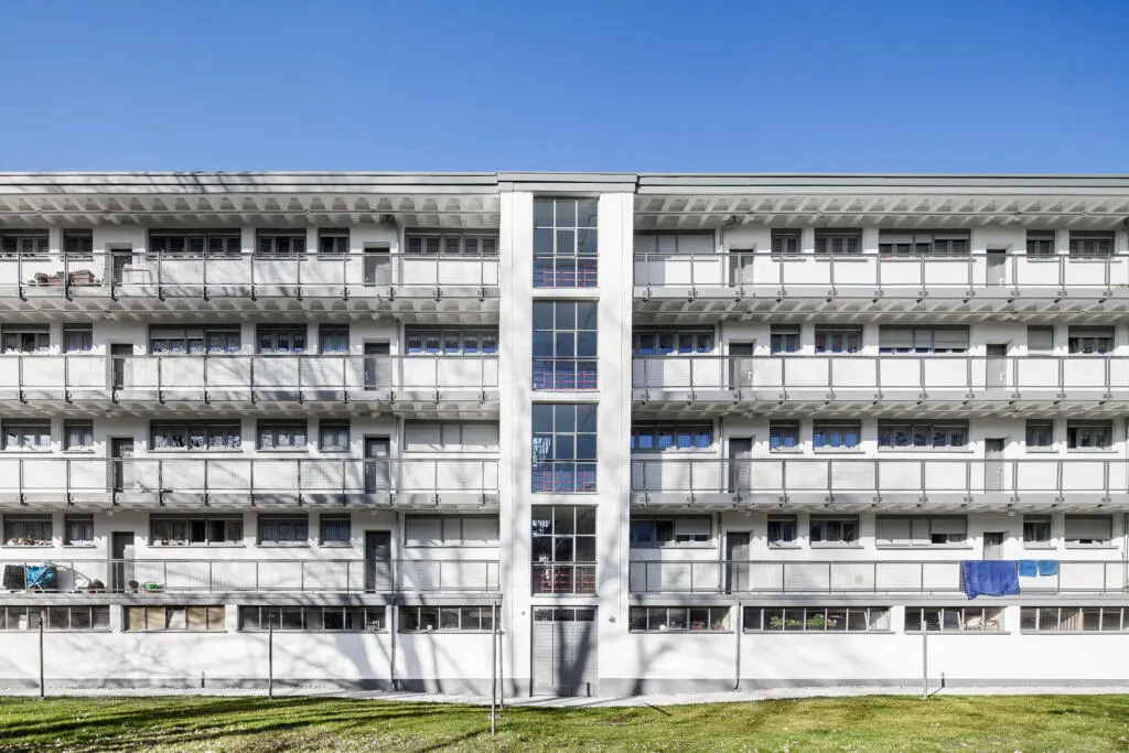 Frontale Ansicht eines weißen, mehrgeschossigen Wohngebäudes im modernistischen Stil mit durchgehenden Laubengängen und zentralem verglasten Treppenhaus unter blauem Himmel.