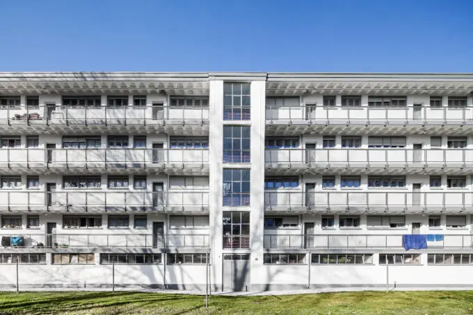 Frontale Ansicht eines weißen, mehrgeschossigen Wohngebäudes im modernistischen Stil mit durchgehenden Laubengängen und zentralem verglasten Treppenhaus unter blauem Himmel.