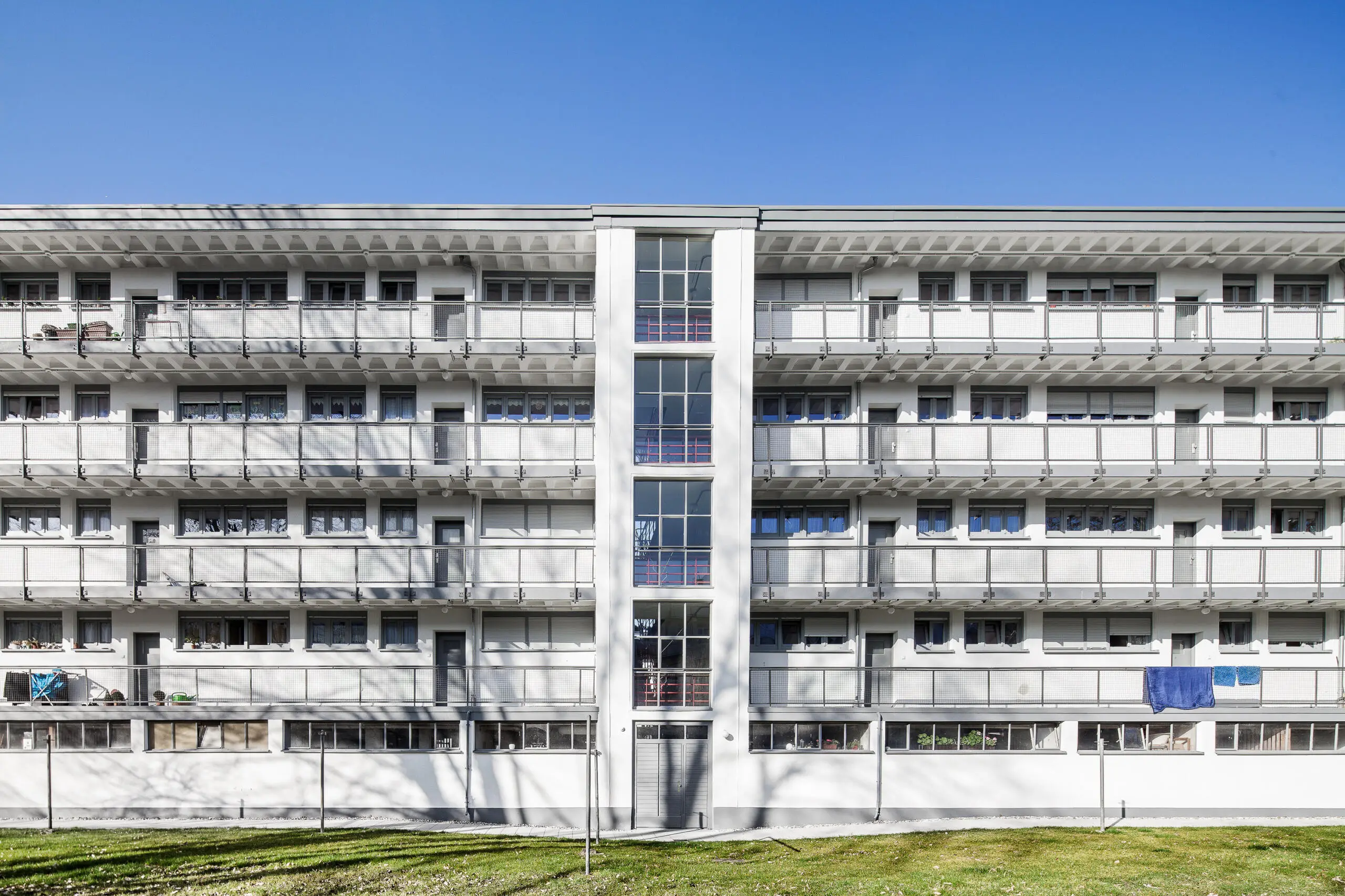 Frontale Ansicht eines weißen, mehrgeschossigen Wohngebäudes im modernistischen Stil mit durchgehenden Laubengängen und zentralem verglasten Treppenhaus unter blauem Himmel.
