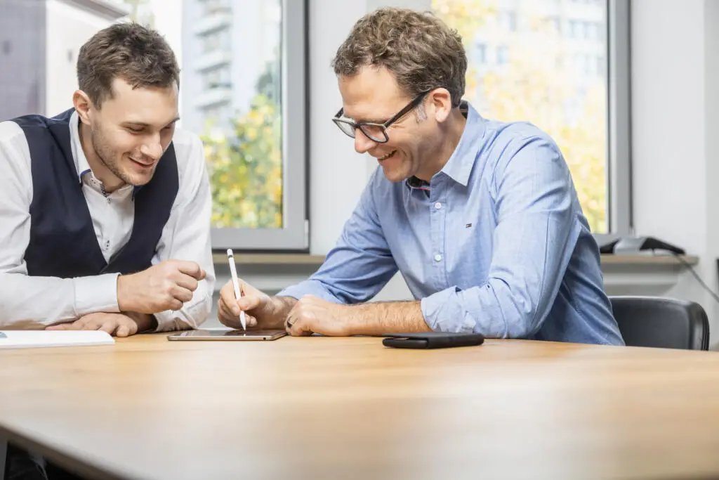 Zwei Männer in Business-Kleidung arbeiten gemeinsam an einem Tablet mit Stylus an einem Holztisch in einem hellen Büro.