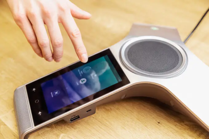 Eine Hand bedient das Touchscreen-Display eines modernen silbernen Konferenztelefons auf einem hellen Holztisch.