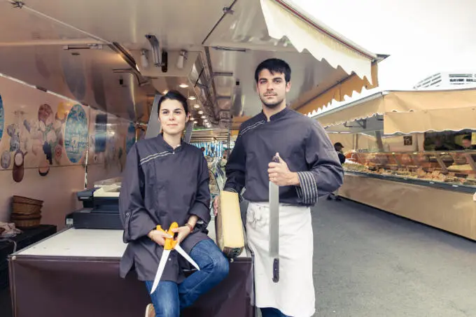 Ein Mann und eine Frau in Arbeitskleidung stehen vor einem mobilen Marktstand für Käse und halten ein Messer sowie eine Schere in den Händen.