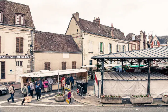 Menschen beim Einkauf an einem weißen Marktstand neben einer überdachten Markthalle in einer historischen Altstadt