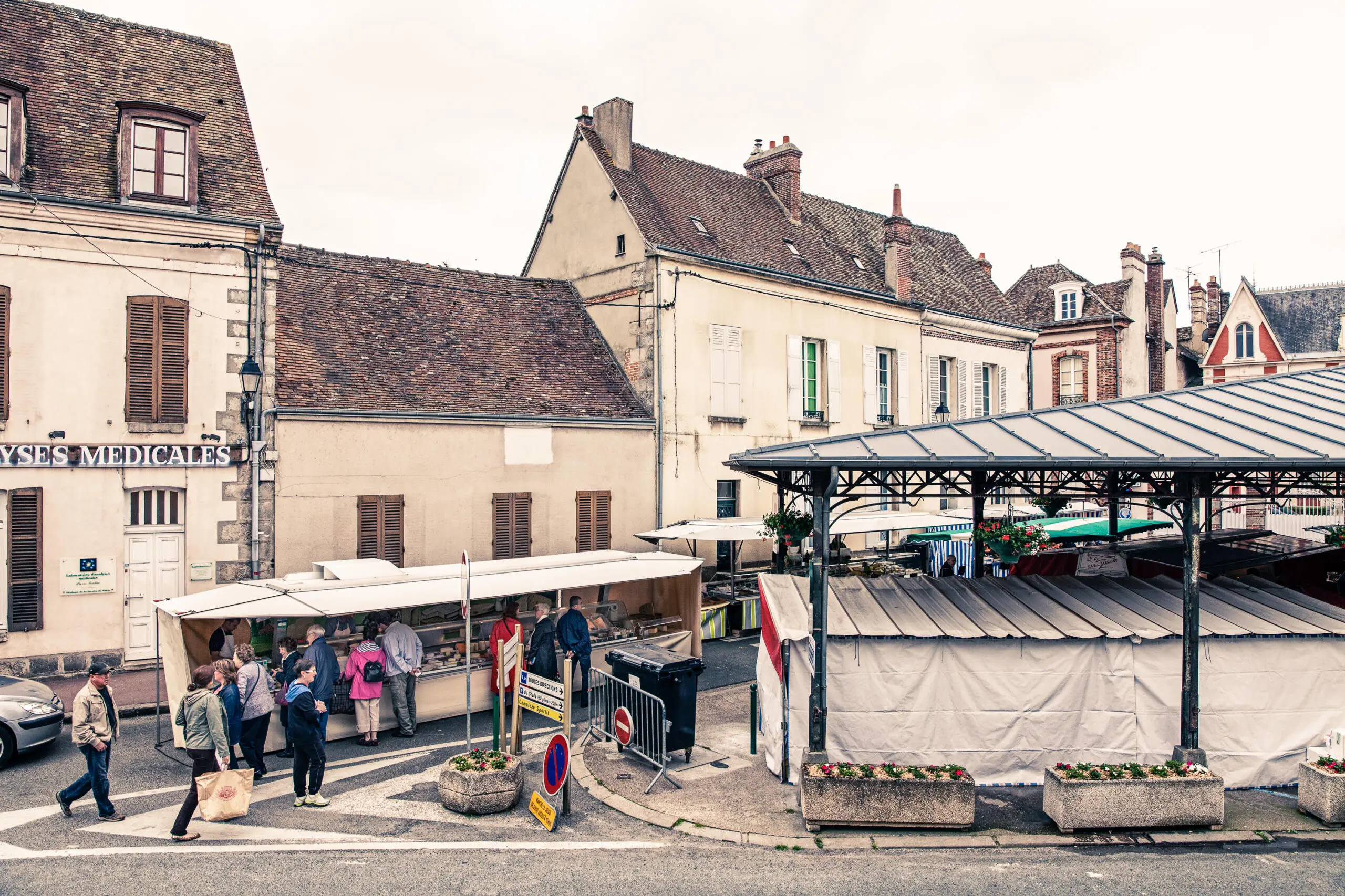 Menschen beim Einkauf an einem weißen Marktstand neben einer überdachten Markthalle in einer historischen Altstadt