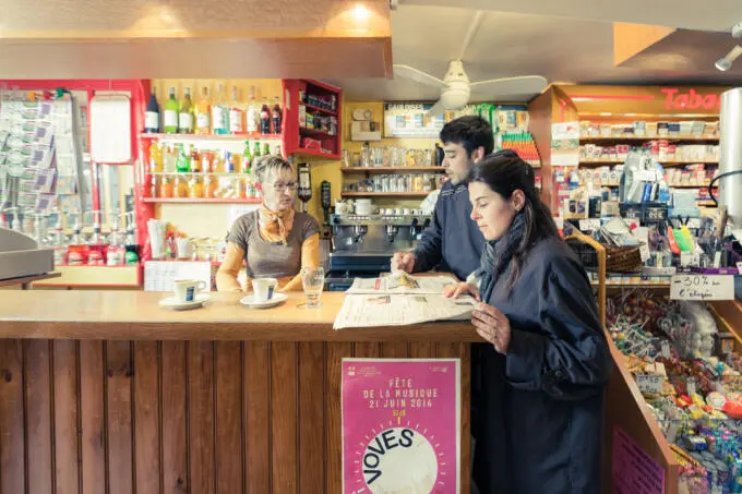 Eine Verkäuferin hinter einem Holztresen im Gespräch mit zwei Kunden, während eine Kundin Zeitung liest und Kaffeetassen auf der Theke stehen
