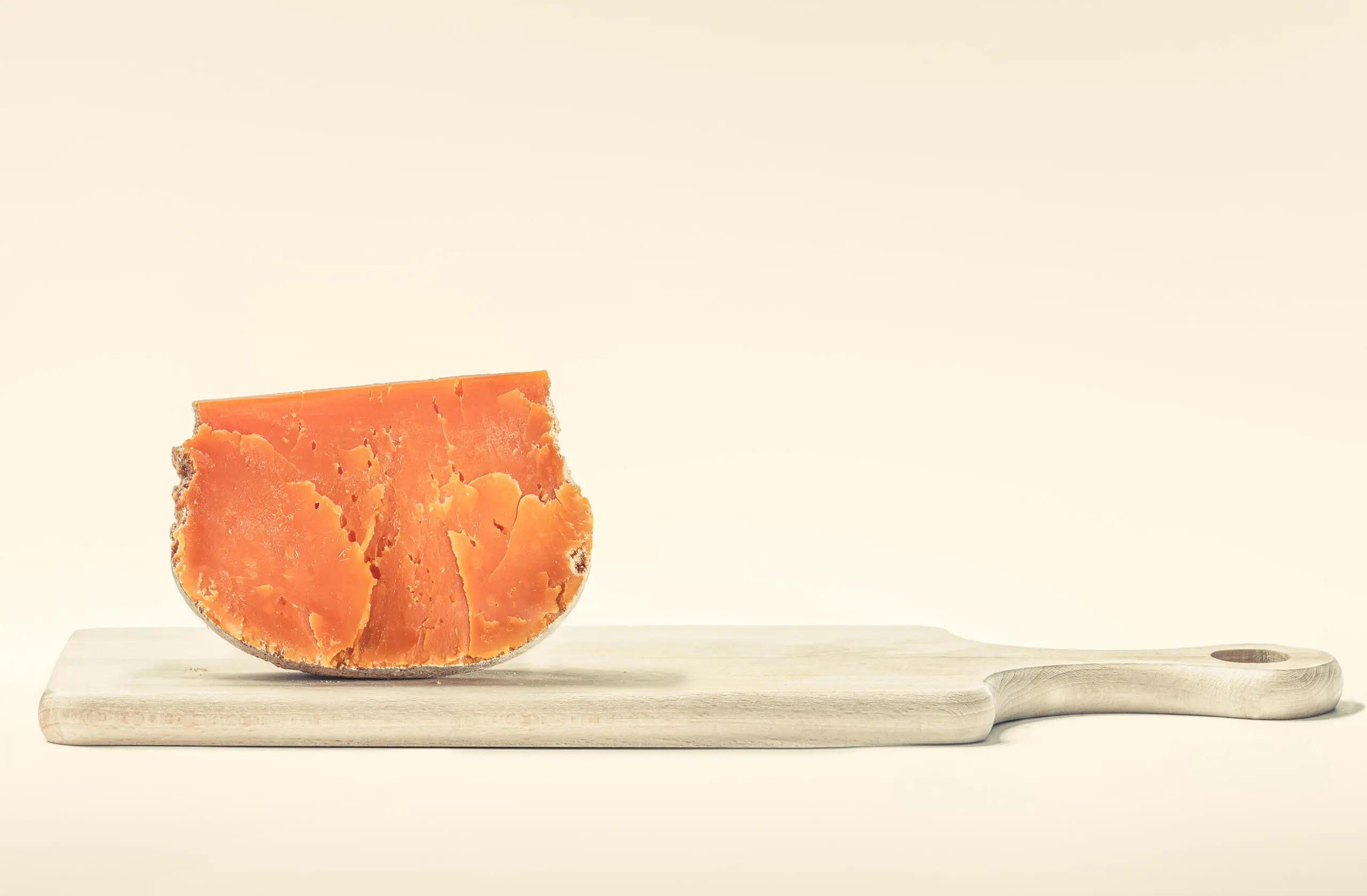 Ein Keil gereifter, intensiv orangefarbener Hartkäse steht aufrecht auf einem hellen Holzschneidebrett vor neutralem Hintergrund.