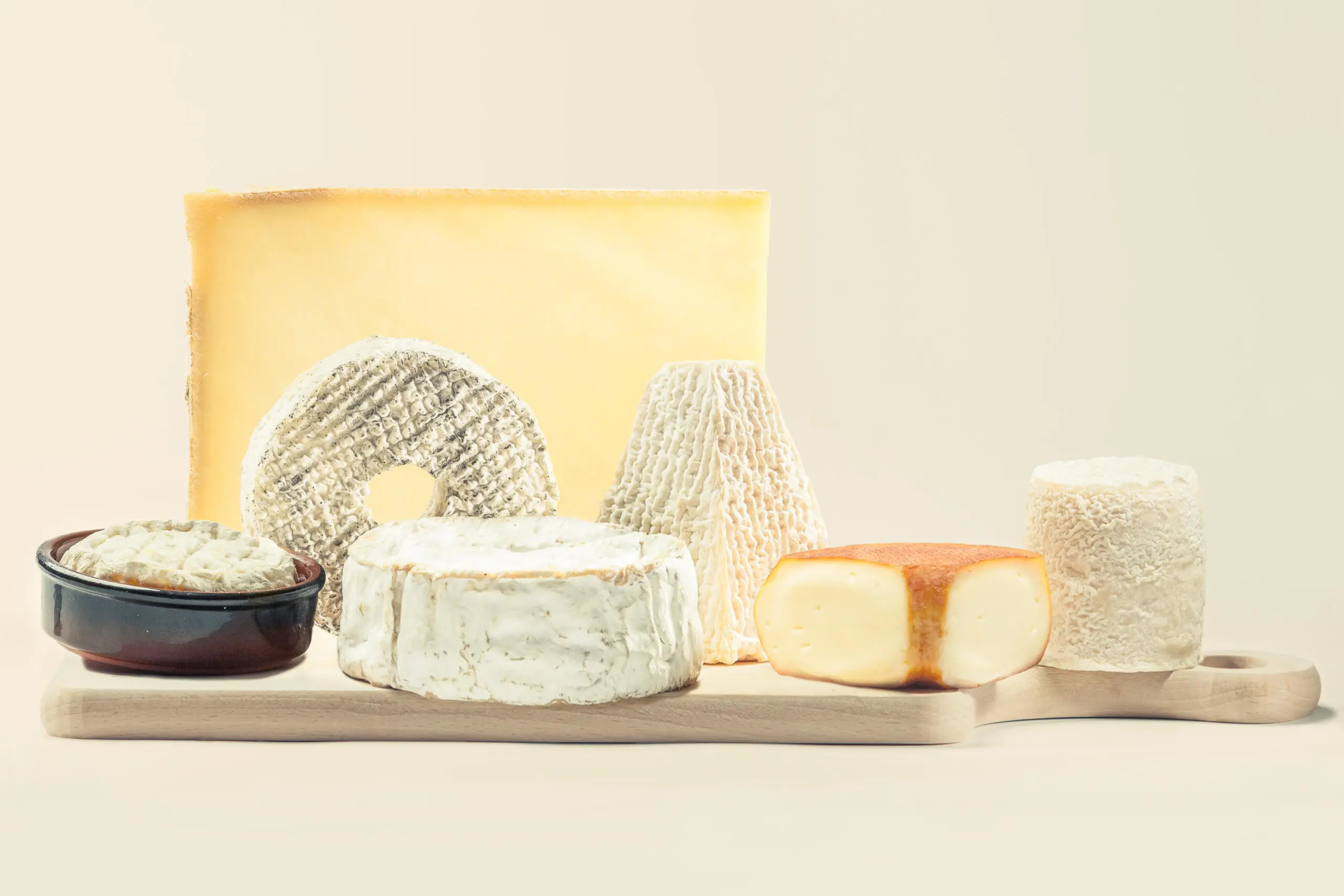 Verschiedene Käsesorten wie Hartkäse, Weichkäse und Ziegenkäse sind auf einem hellen Holzbrett vor neutralem Hintergrund arrangiert.