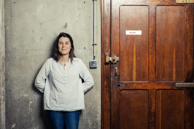 Eine lächelnde Frau in weißem Hemd steht vor einer grauen Betonwand neben einer braunen Holztür mit der Aufschrift Privé.
