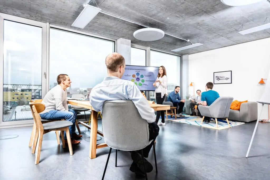 Eine Mitarbeiterin hält eine Präsentation an einem digitalen Bildschirm vor einer Gruppe von Kollegen in einem hellen Büro mit Sichtbetondecke und großen Fenstern.