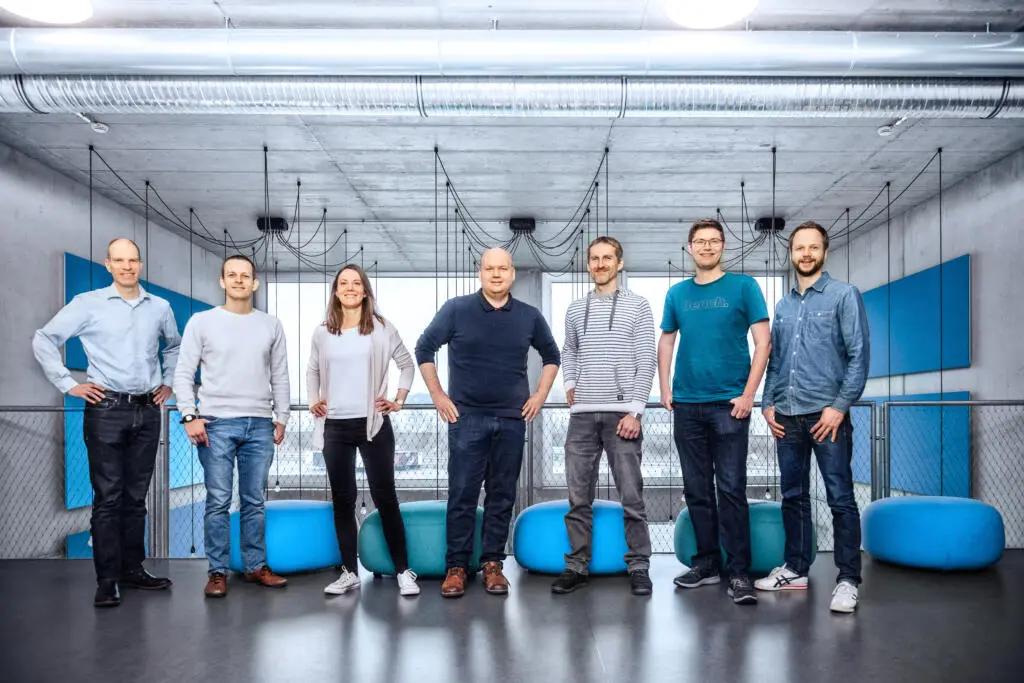 Sieben Mitarbeiter stehen nebeneinander in einem modernen Büro mit Sichtbetondecke, sichtbaren Lüftungsrohren und blauen Akustikelementen.