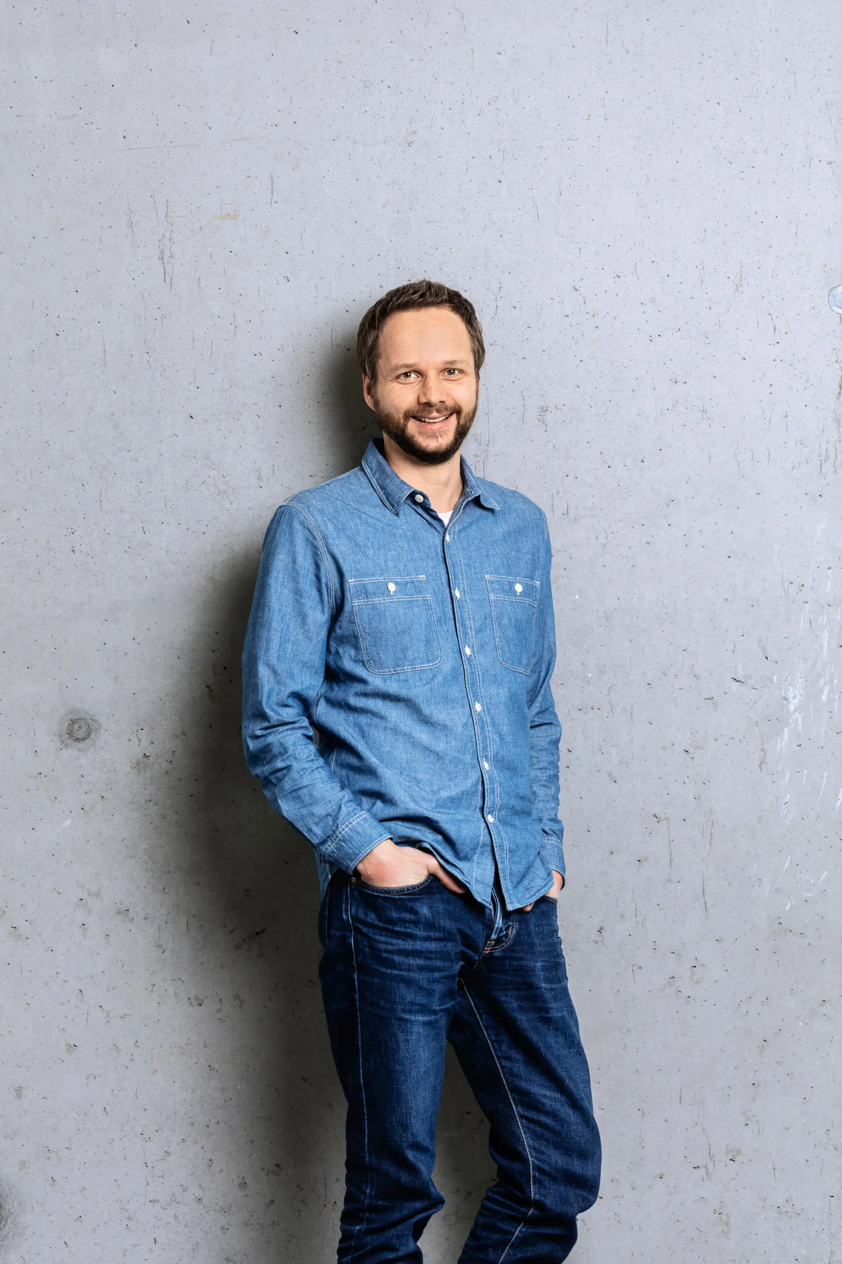 Ein lächelnder Mann mit Bart in Jeanshemd und Jeanshose steht mit Händen in den Taschen vor einer grauen Sichtbetonwand.