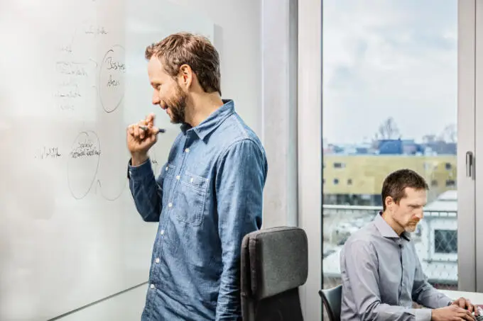 Ein stehender Mann im Jeanshemd betrachtet lächelnd Notizen an einem Whiteboard, während ein Kollege im Hintergrund am Schreibtisch vor einem Fenster arbeitet.
