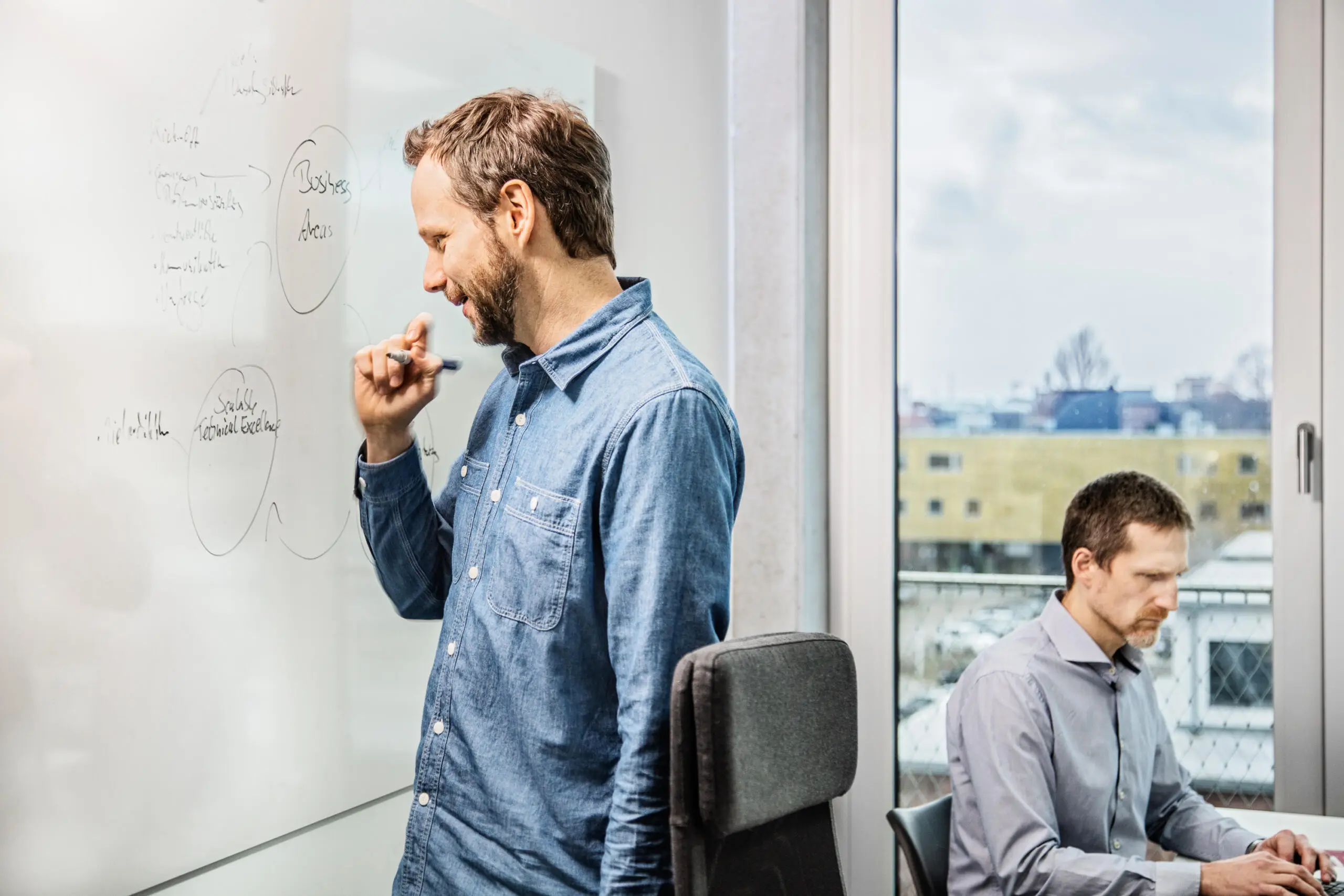 Ein stehender Mann im Jeanshemd betrachtet lächelnd Notizen an einem Whiteboard, während ein Kollege im Hintergrund am Schreibtisch vor einem Fenster arbeitet.