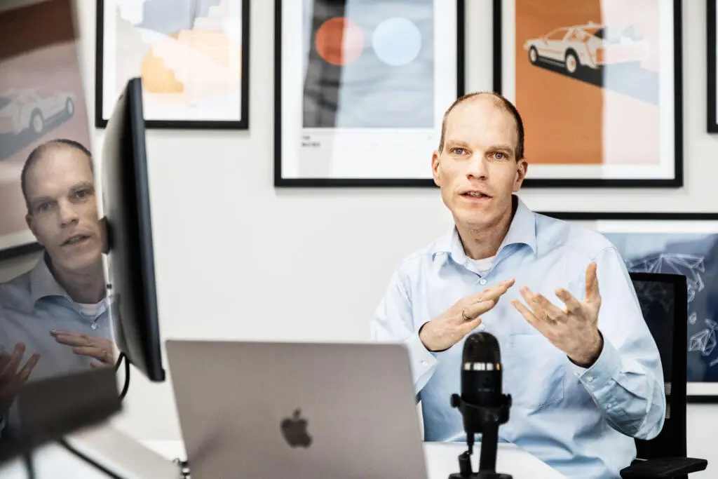 Ein Mann im hellblauen Hemd sitzt an einem Schreibtisch mit Laptop und Mikrofon und gestikuliert während des Sprechens.