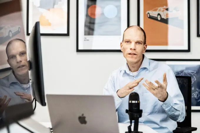 Ein Mann im hellblauen Hemd sitzt an einem Schreibtisch mit Laptop und Mikrofon und gestikuliert während des Sprechens.