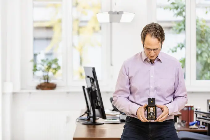 Ein Mann steht in einem hellen Büro und blickt konzentriert auf eine historische zweiäugige Spiegelreflexkamera in seinen Händen.