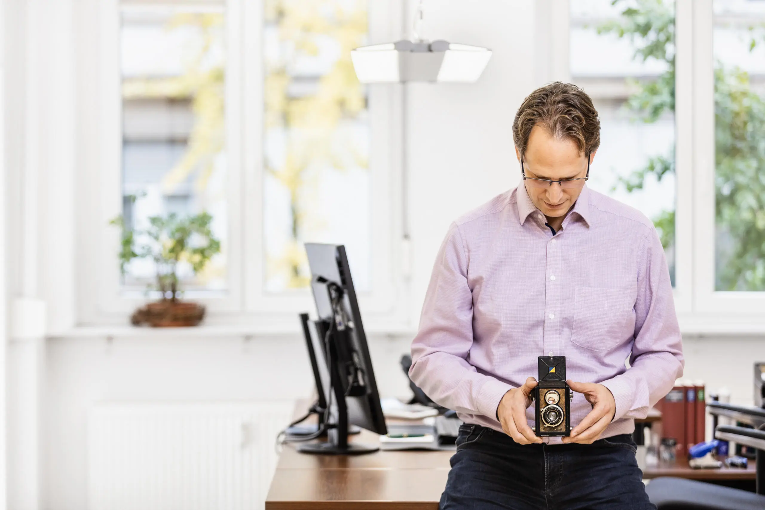 Ein Mann steht in einem hellen Büro und blickt konzentriert auf eine historische zweiäugige Spiegelreflexkamera in seinen Händen.