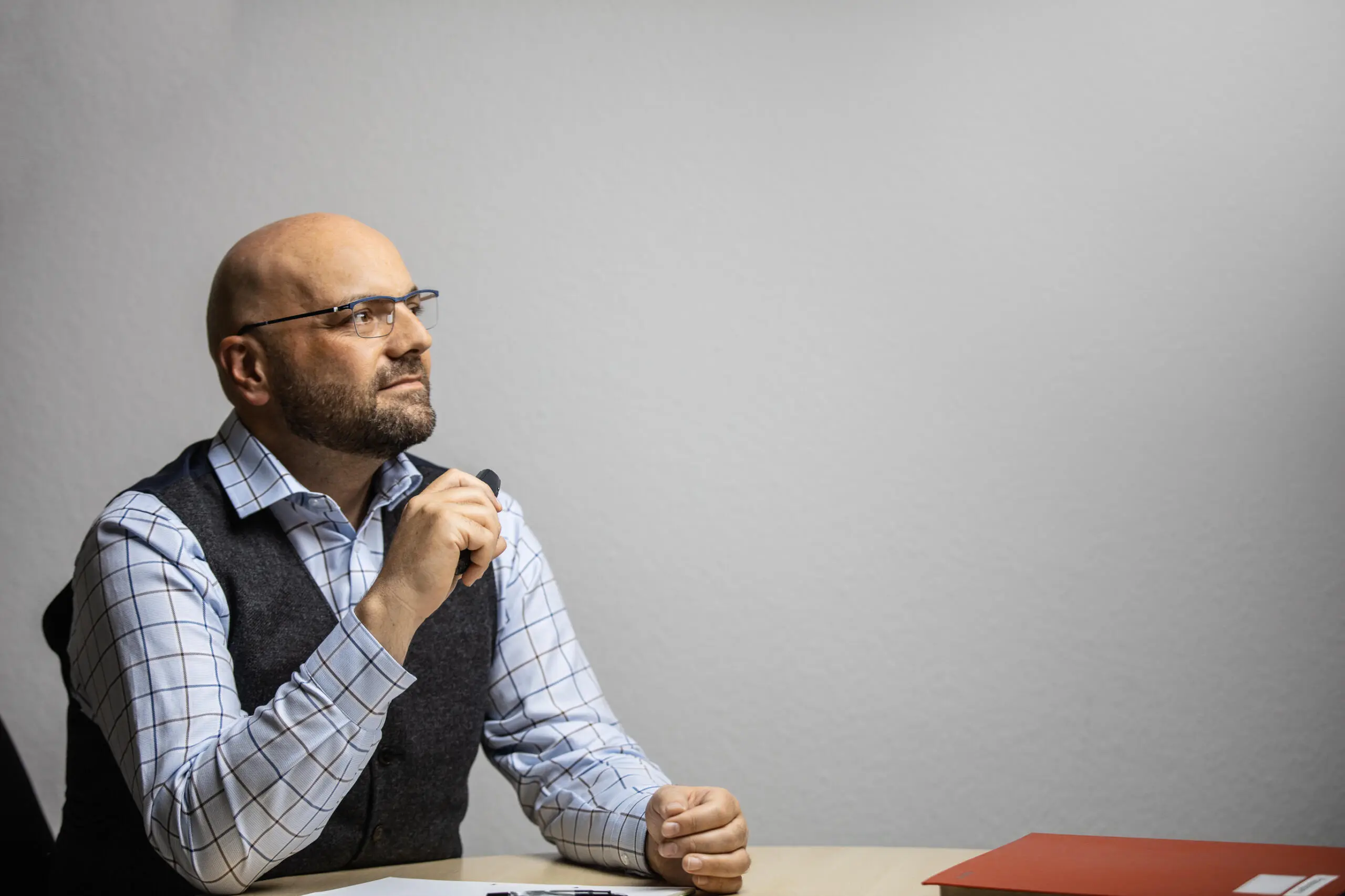 Ein Mann mit Brille und Weste sitzt nachdenklich mit einem Stift in der Hand an einem Schreibtisch vor grauem Hintergrund.