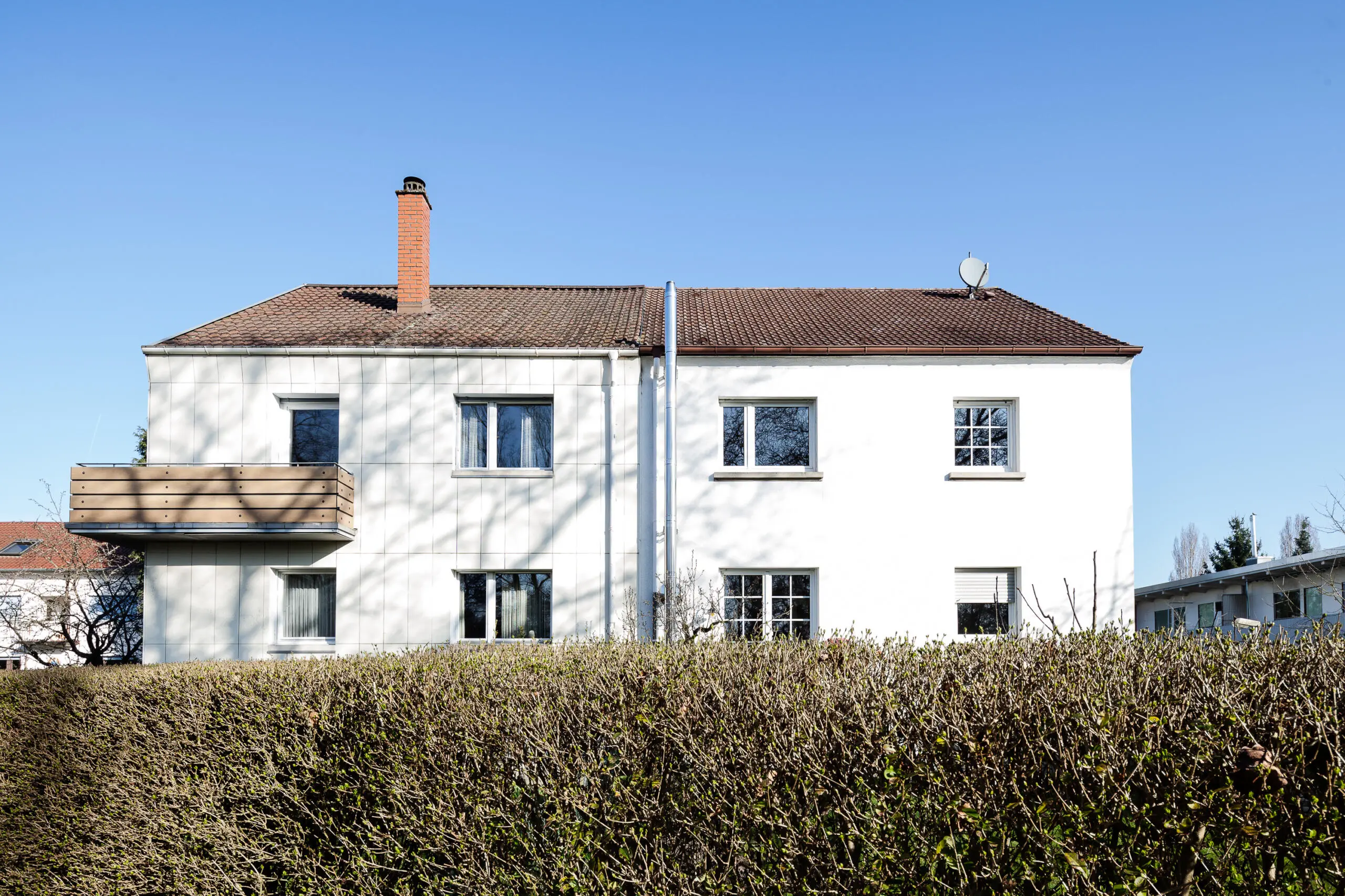 Frontalansicht eines zweigeschossigen weißen Wohnhauses mit Satteldach und Holzbalkon hinter einer dichten Hecke unter blauem Himmel.