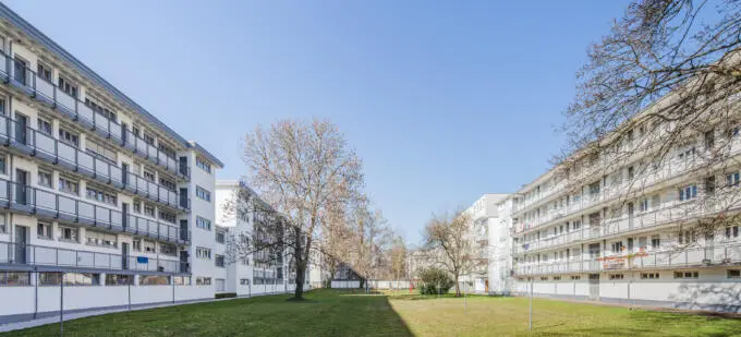 Grüner Innenhof zwischen zwei weißen mehrstöckigen Wohnriegeln der Nachkriegsmoderne mit Balkonen und Bäumen unter blauem Himmel.
