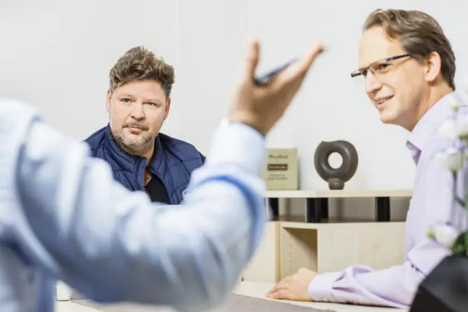 Zwei Männer im Gespräch mit einem unscharf im Vordergrund sichtbaren Kollegen in einem hellen Büro