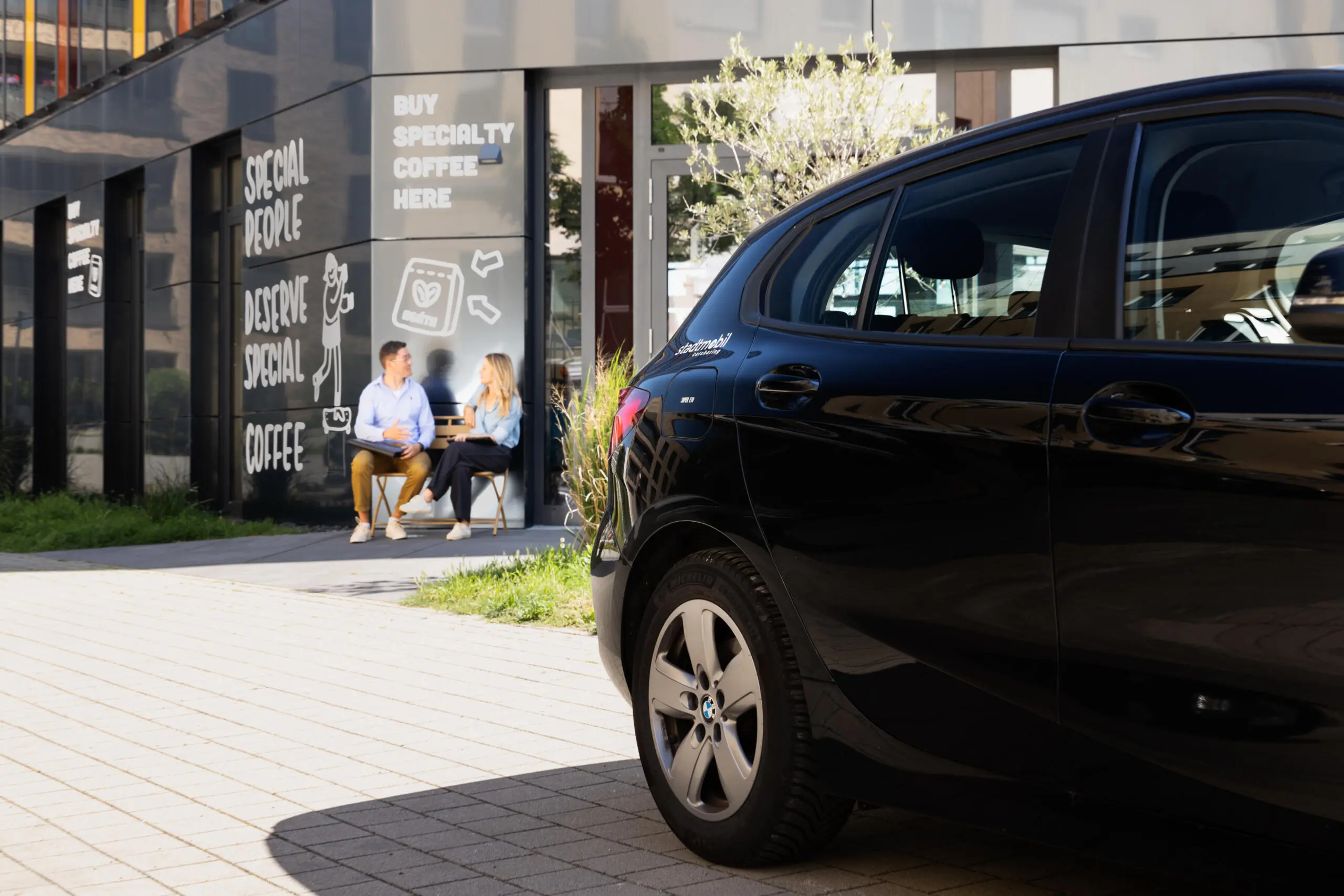 Heckansicht eines schwarzen Fahrzeugs im Vordergrund vor einem modernen Café mit zwei sitzenden Personen im Gespräch