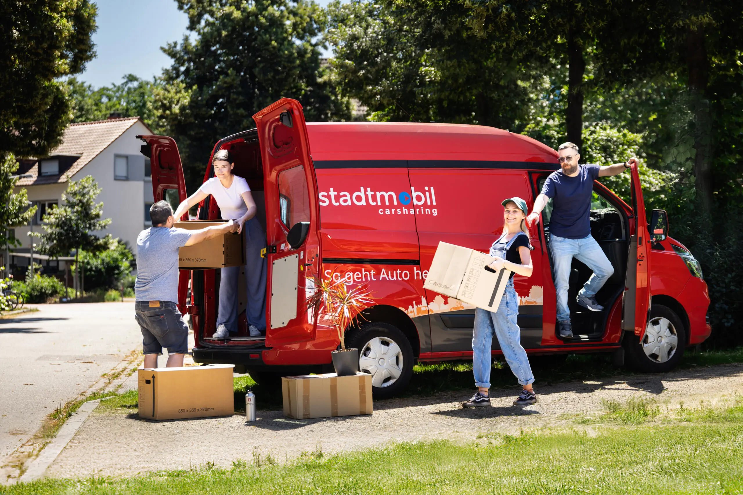 Group of people loading cardboard boxes into a red carsharing transporter van on a sunny residential street