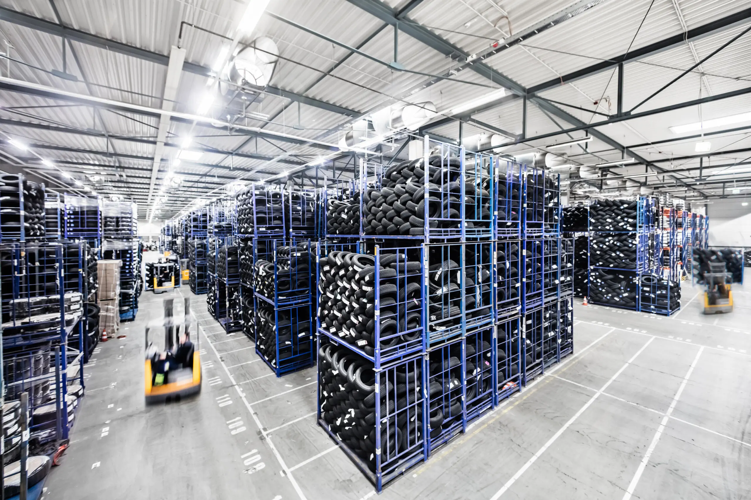 Weitwinkelaufnahme einer großen Lagerhalle mit hohen Regalen voller Autoreifen und einem fahrenden Gabelstapler mit Bewegungsunschärfe