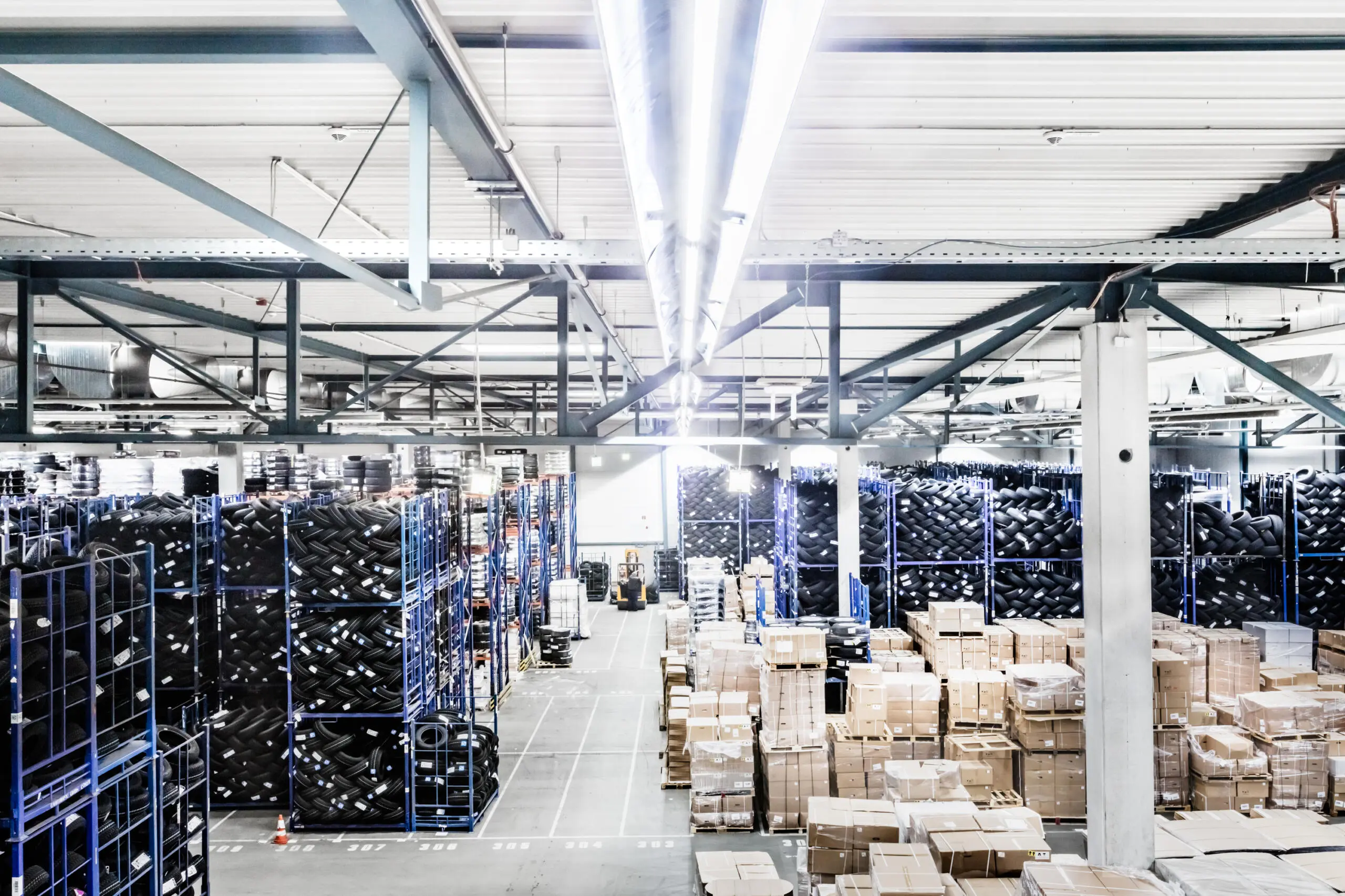 Innenansicht einer großen Lagerhalle mit blauen Regalen voller Reifen links und Paletten mit Kartons rechts unter heller Deckenbeleuchtung