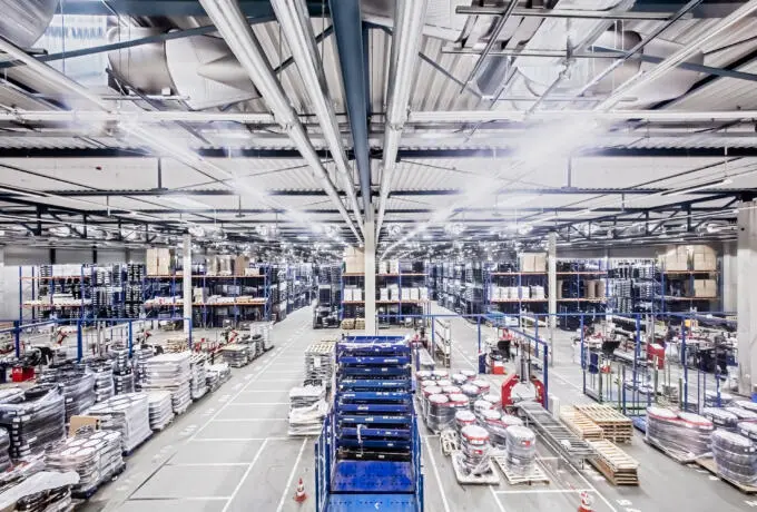 Weitwinkelaufnahme einer hell erleuchteten Lagerhalle mit Hochregalen, Lüftungsrohren an der Decke, Paletten mit Waren und blauen Transportwagen im Mittelgang