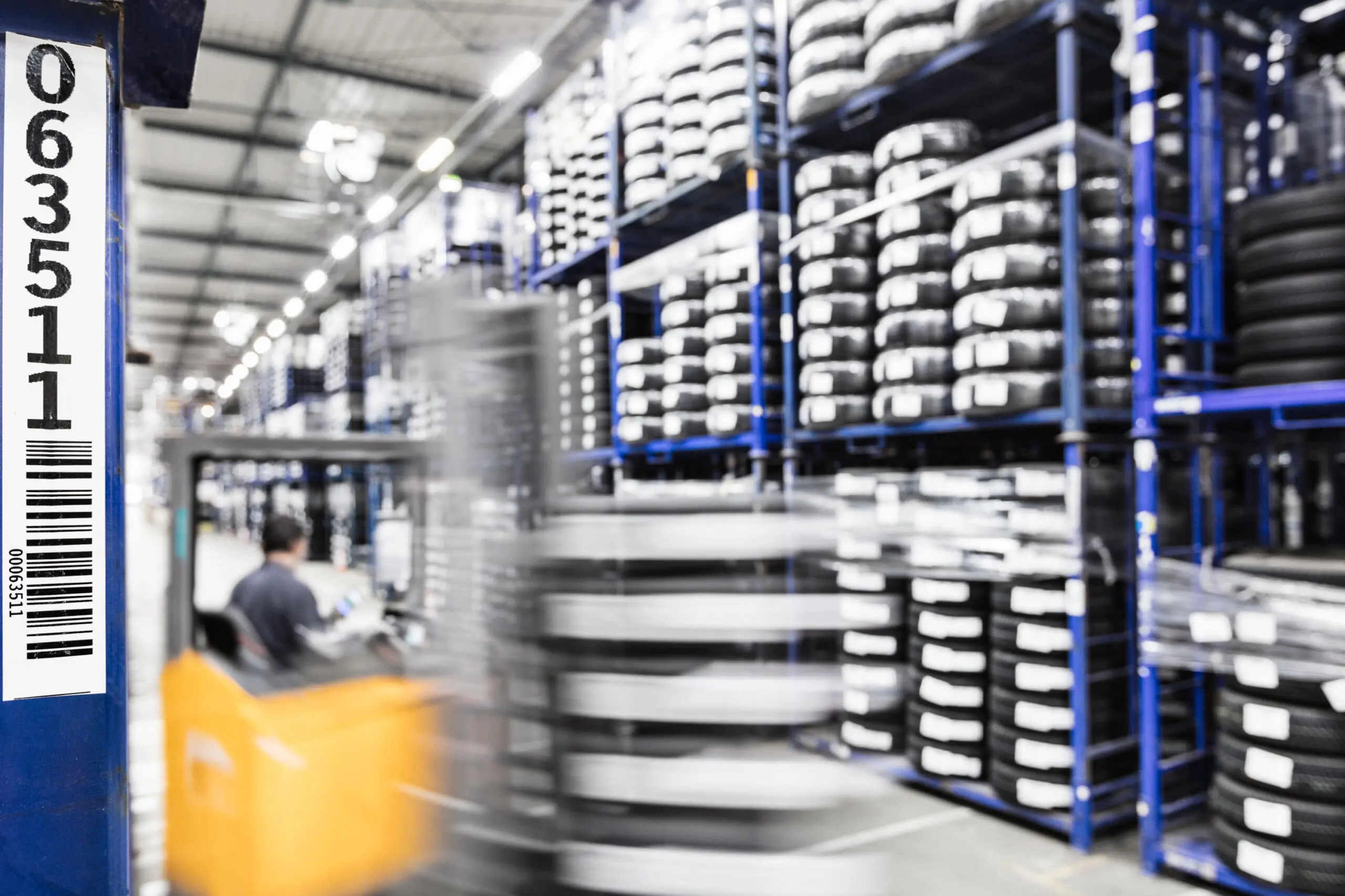 Gabelstapler fährt durch Reifenlager mit Bewegungsunschärfe, im Vordergrund scharfe Regalnummerierung mit Barcode